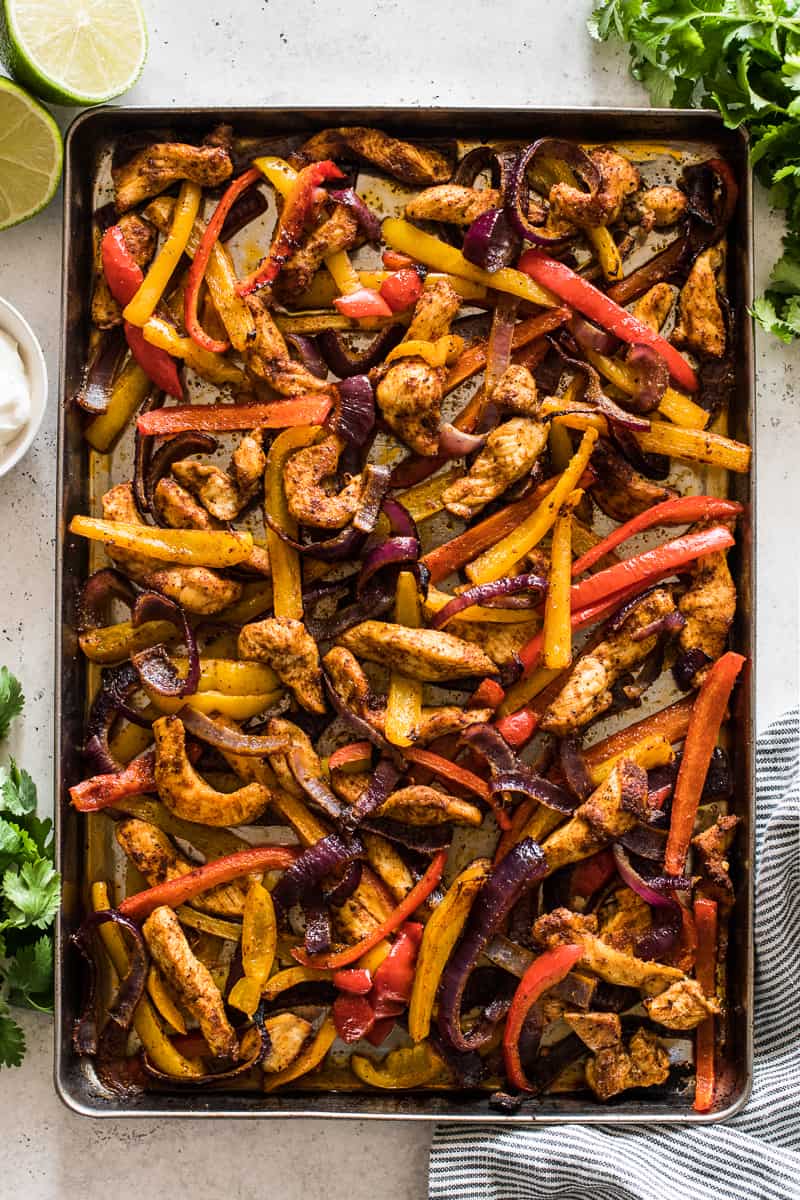 Sheet pan chicken fajitas on a table with cilantro, sour cream, and lime juice.
