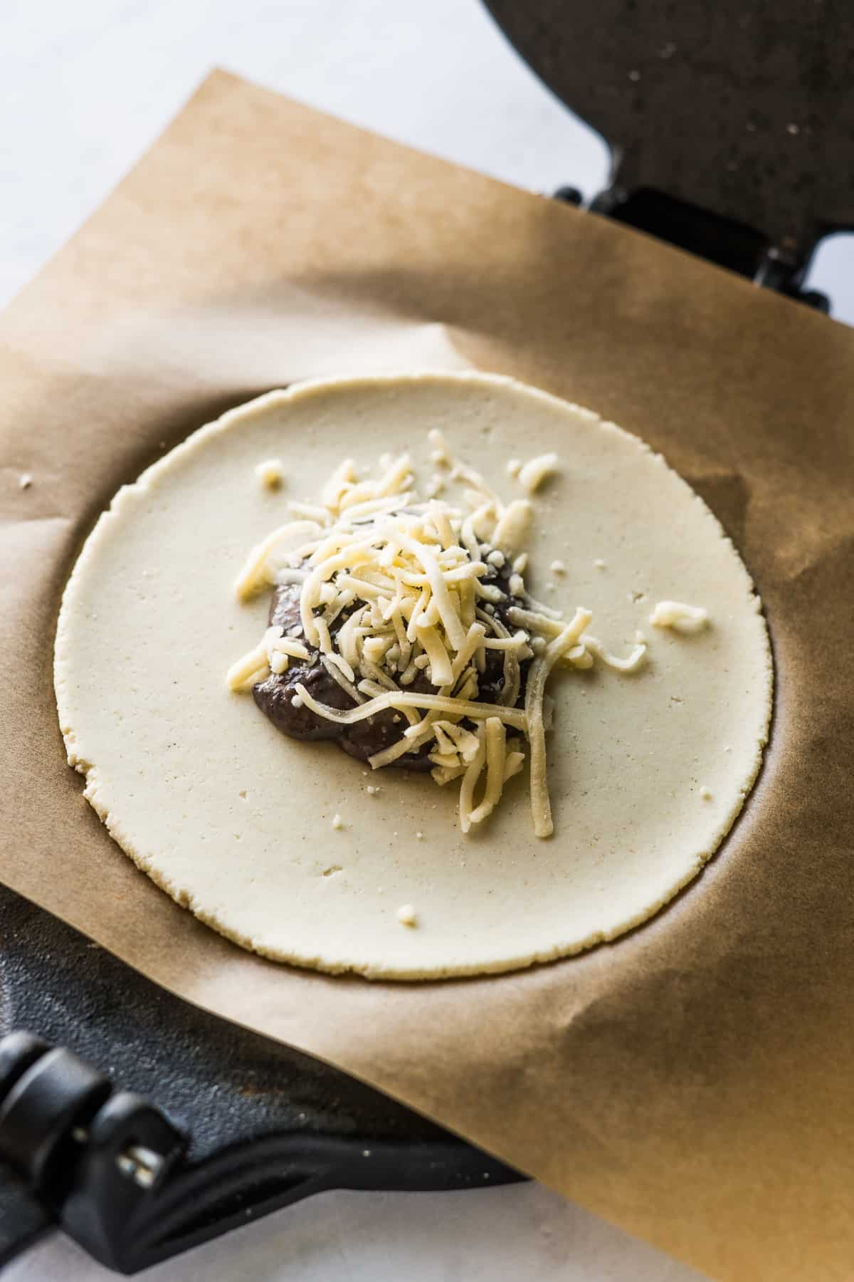 Pressed masa harina on a tortilla press topped with black beans and cheese.
