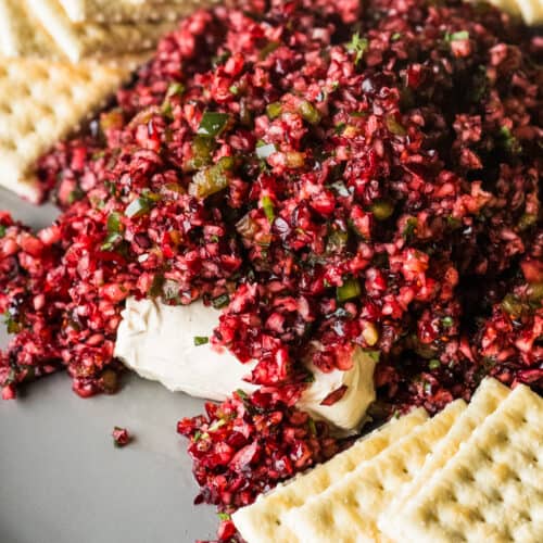 Cranberry salsa on a serving plate on top of a block of cream cheese with some crackers on the side.