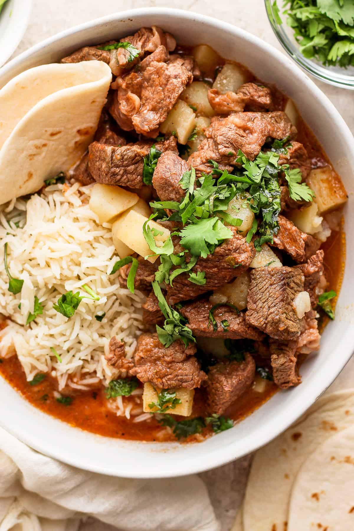 A bowl of carne con papas served with rice and flour tortillas.