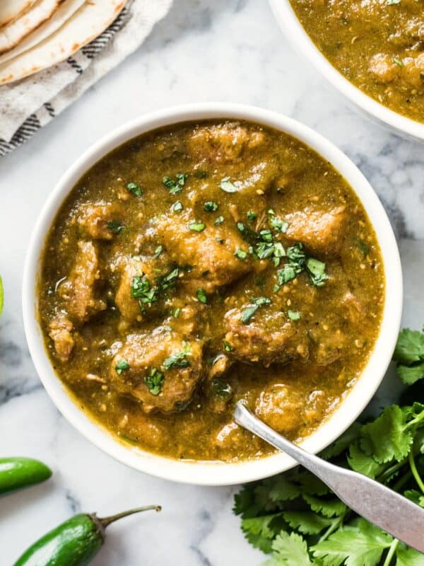 Pork chile verde in a bowl served over in a bowl with fresh cilantro and lime wedges and flour tortillas on the side.