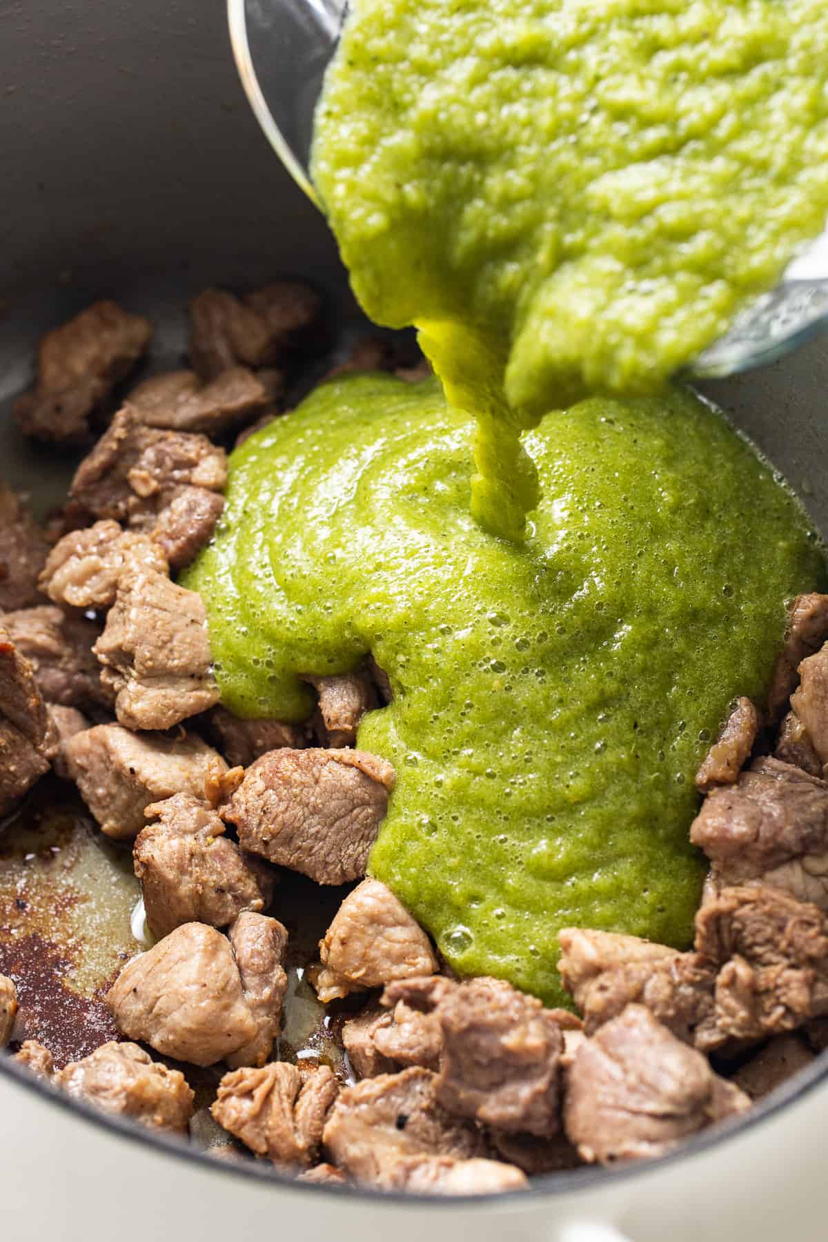Blended salsa verde being poured into a pot of seared pork shoulder chunks.