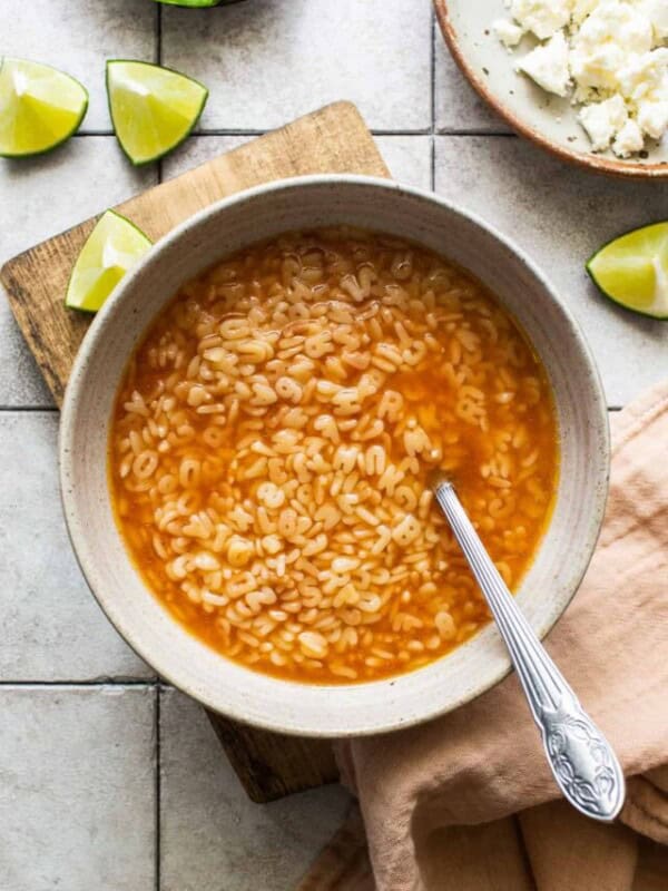 A bowl of sopa de letras on a table with limes, cheese, and avocado on the side.