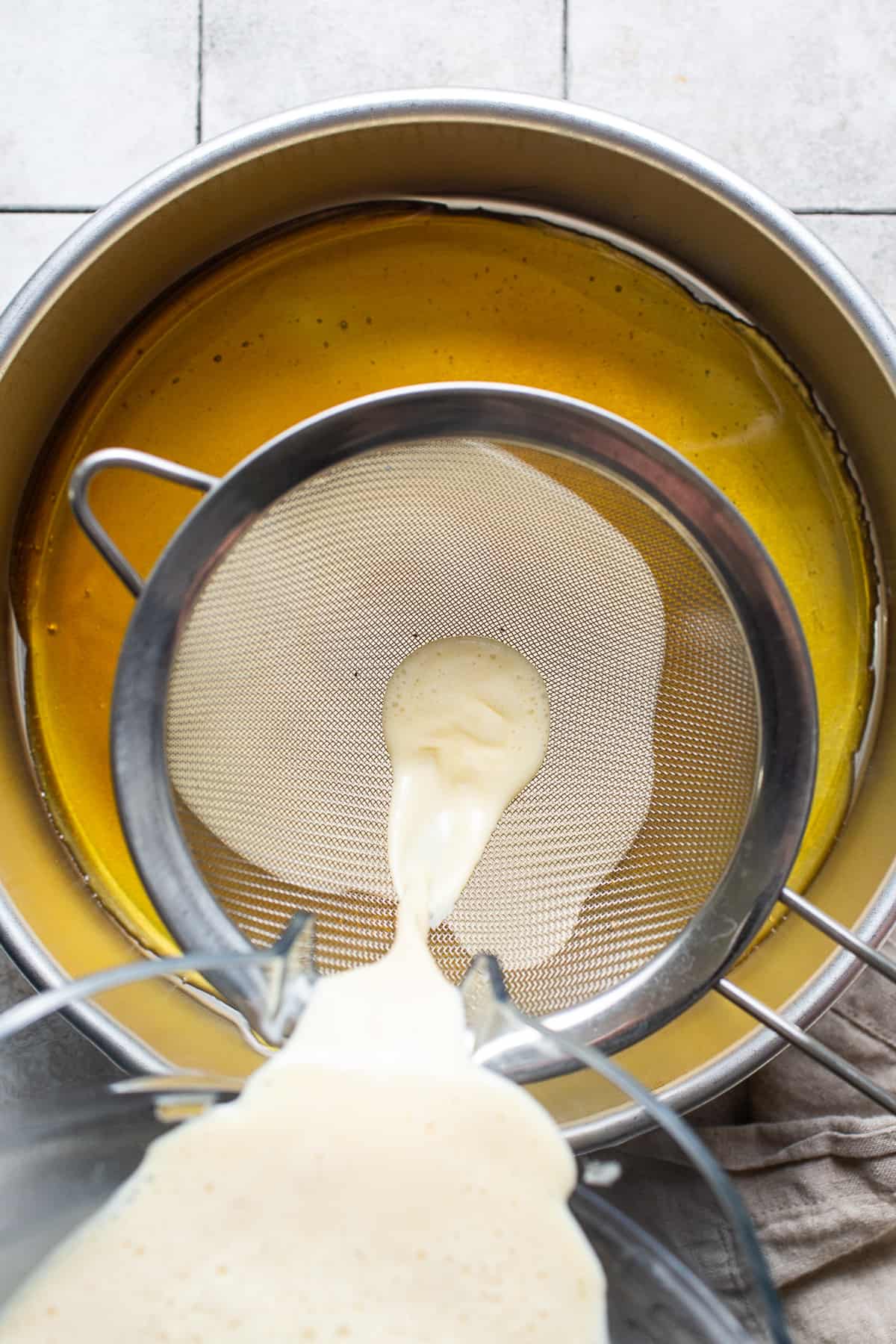 The blended flan de queso mixture being poured through a fine mesh strainer and into a round cake pan.