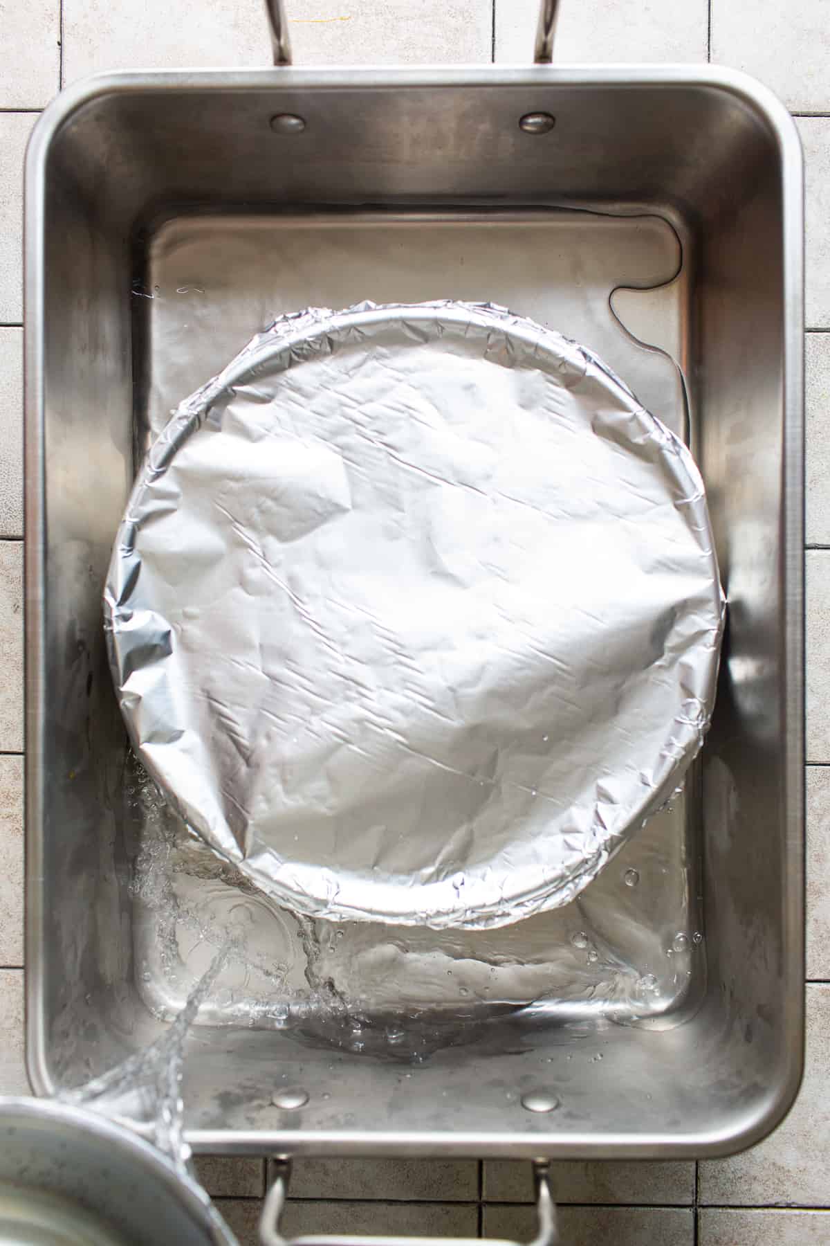 A round cake pan covered with aluminum foil with water being poured into a large pan to create a baine marie or water bath.
