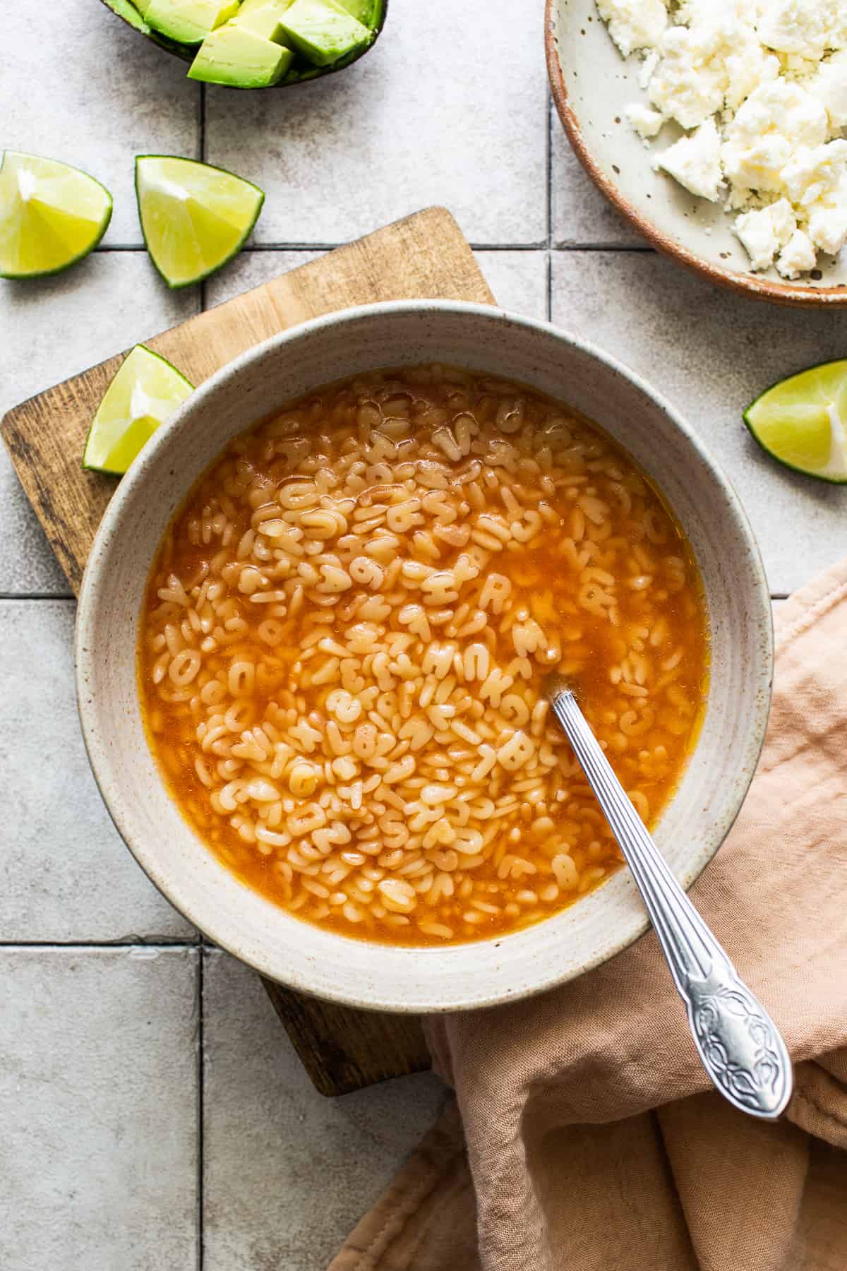 A bowl of sopa de letras on a table with limes, cheese, and avocado on the side.