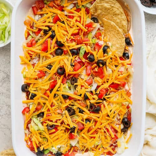 Taco dip in a baking dish topped with shredded cheese, tomatoes, and olive served with tortilla chips.