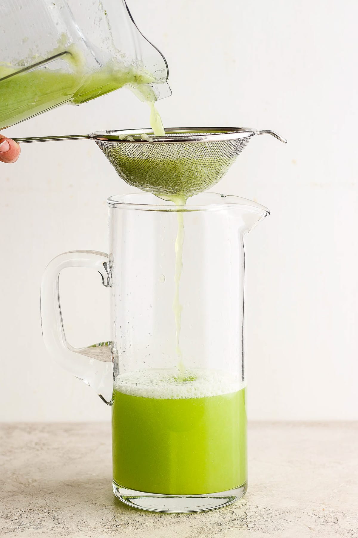 Agua de pepino being strained into a large clear glass pitcher.