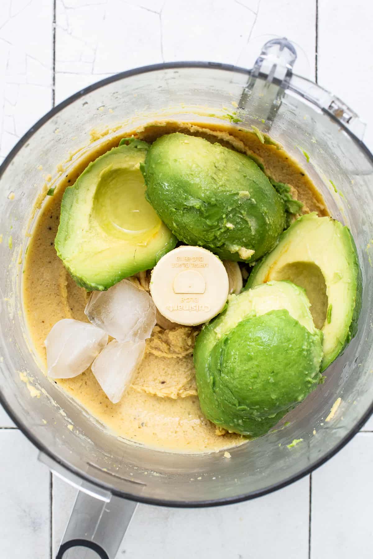 Two avocados, lime juice, and ice cubes in a food processor with blended chickpeas.