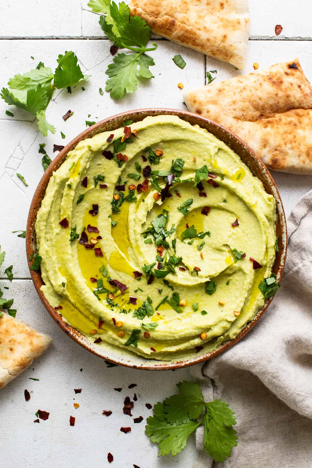 Avocado hummus in a ceramic serving bowl drizzled with olive oil and garnished with chopped cilantro and red pepper flakes.
