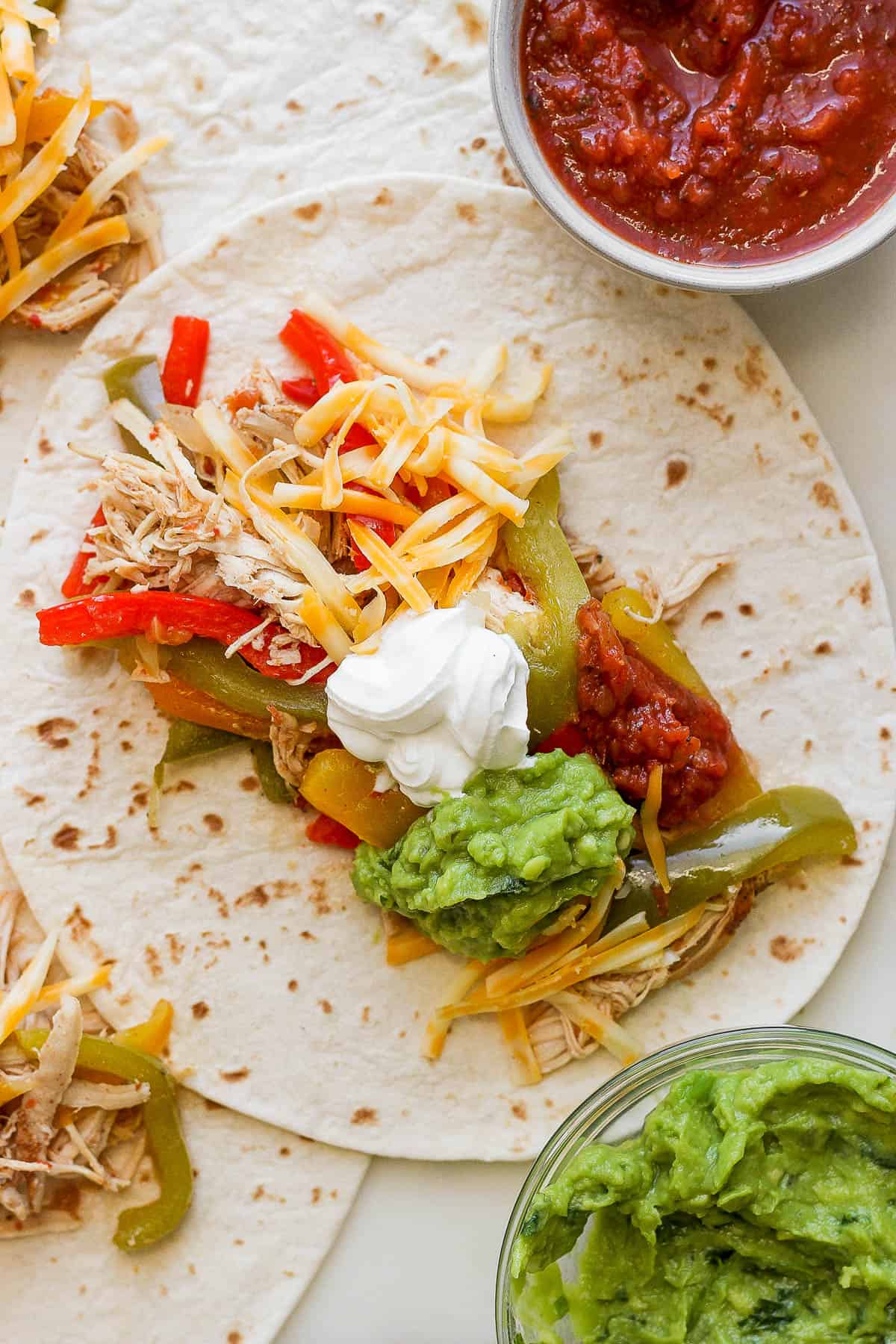 Crockpot chicken fajitas on flour tortillas topped with shredded cheese, guacamole, sour cream, and chunky red salsa.