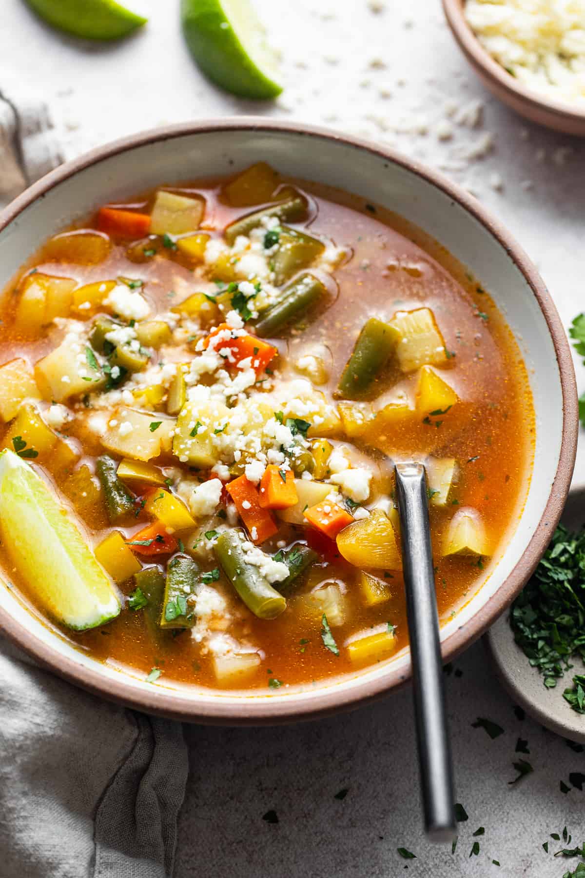 A warm bowl of Mexican vegetable soup served with a lime wedge and sprinkled with cotija cheese.