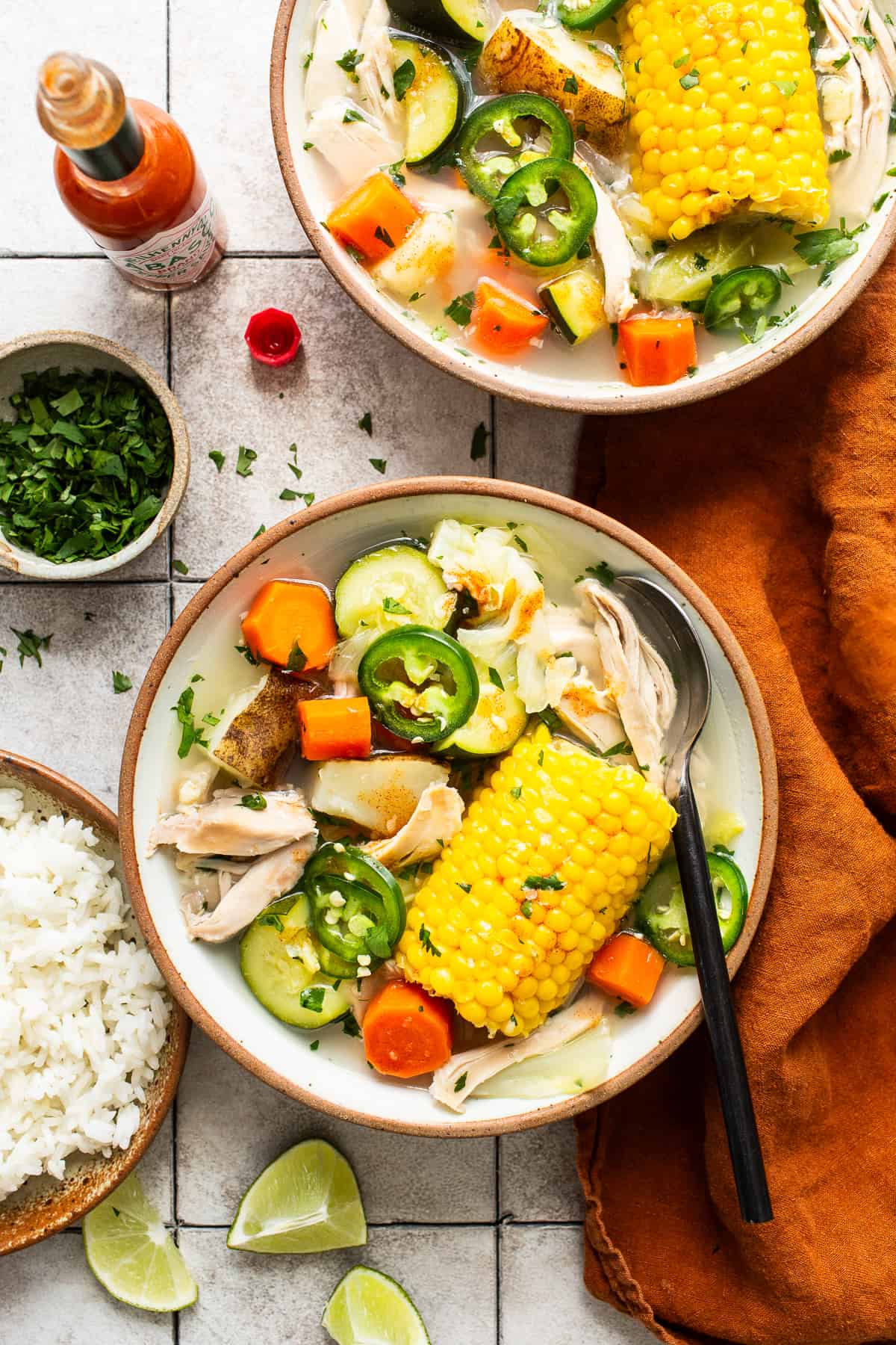 Caldo de pollo (Mexican chicken soup) in two bowls on a table next to white rice, chopped cilantro, and tabasco sauce.