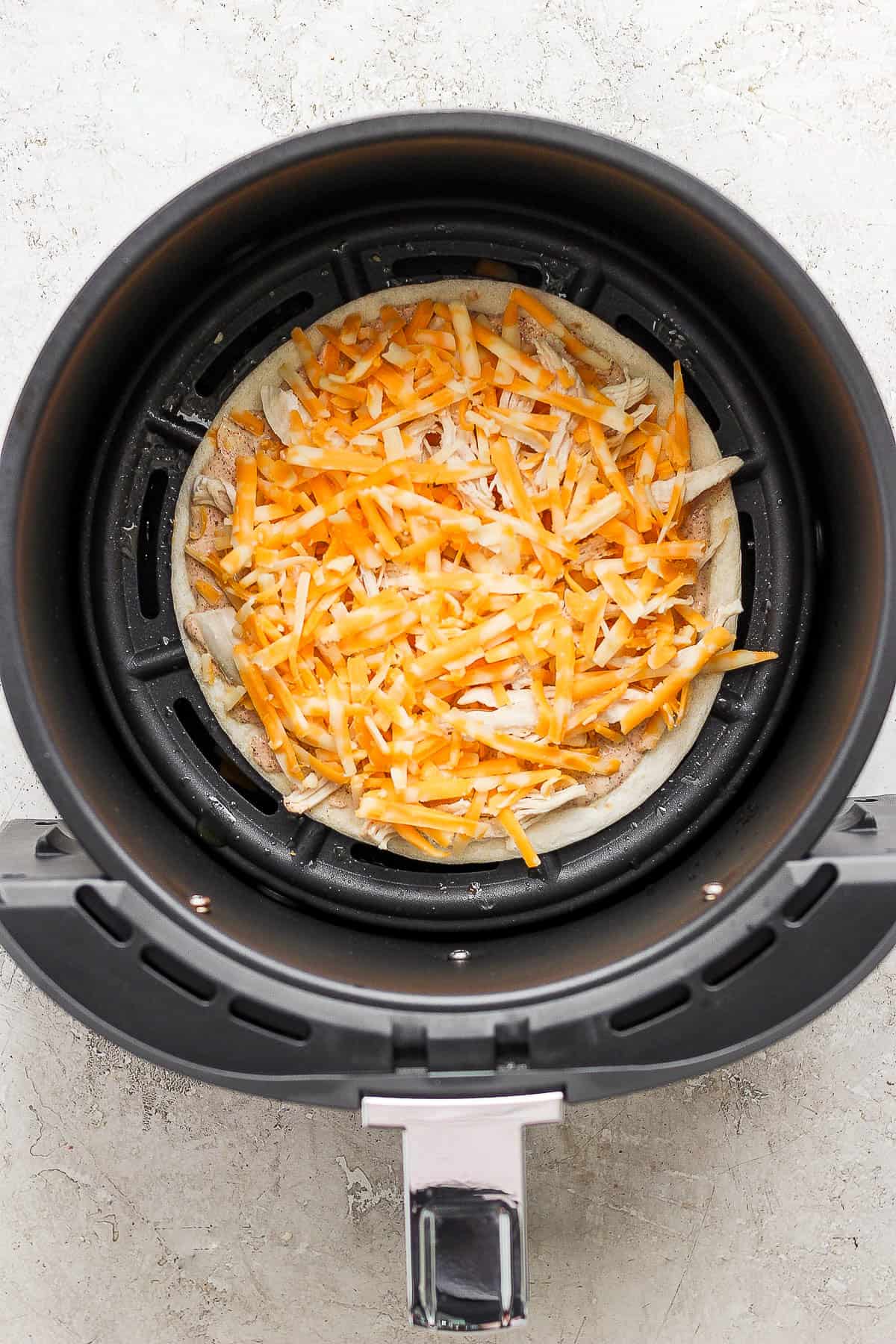 Shredded chicken, shredded cheese, and a chili lime sauce on a flour tortilla sitting in an air fryer for quesadillas.