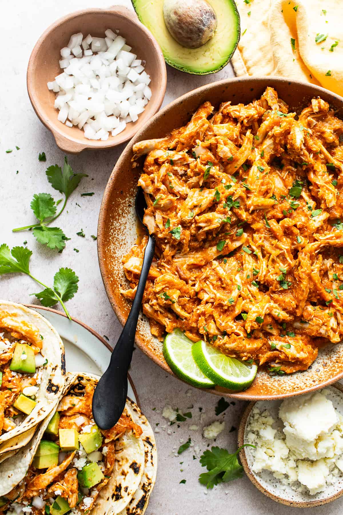 Chicken tinga in a serving bowl alongside chicken tinga tacos, diced onions, queso fresco, avocado, and cilantro.