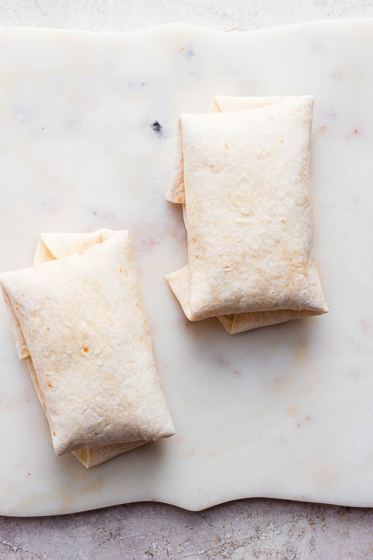 Two folded chimichangas on a cutting board before being cooked.