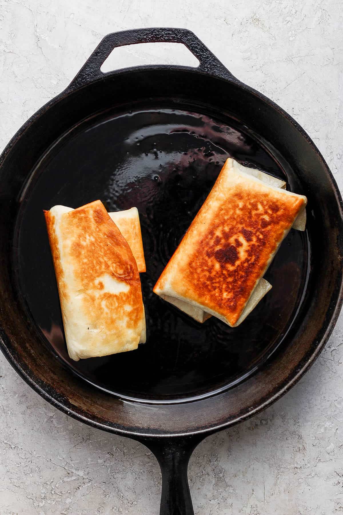 Two chimichangas being pan fried in a skillet with golden brown tops.