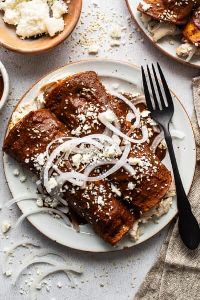 Enmoladas (Chicken Mole Enchiladas) on a plate topped with sesame seeds, queso fresco, and sliced white onions.