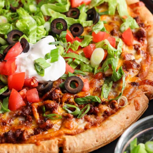 Taco pizza topped with shredded lettuce, sour cream, diced tomatoes, and olives.