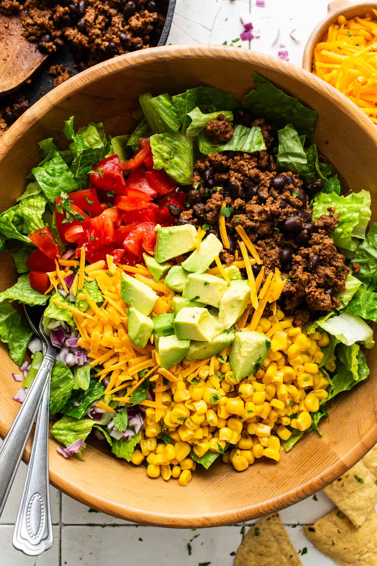 A large bowl filled with shredded lettuce, diced tomatoes, ground beef, black beans, corn, diced red onion, shredded cheese, and diced avocado.