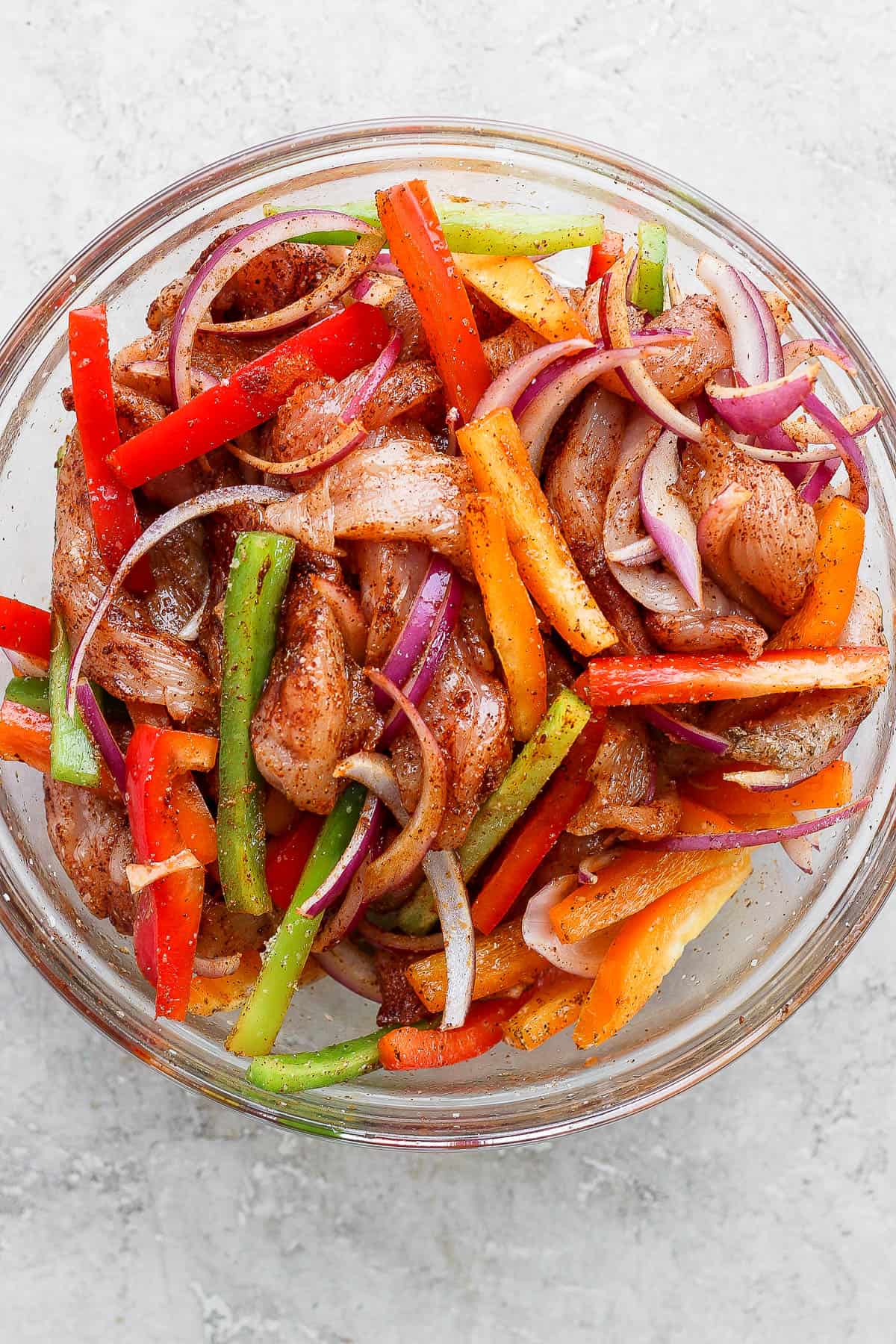 Chicken, vegetables, and seasoning mixed in a clear glass bowl for air fryer chicken fajitas