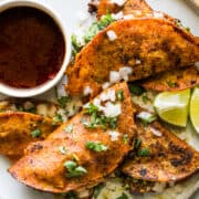 Birria tacos on a plate served with birria consomé and topped with diced white onion, cilantro, and limes.