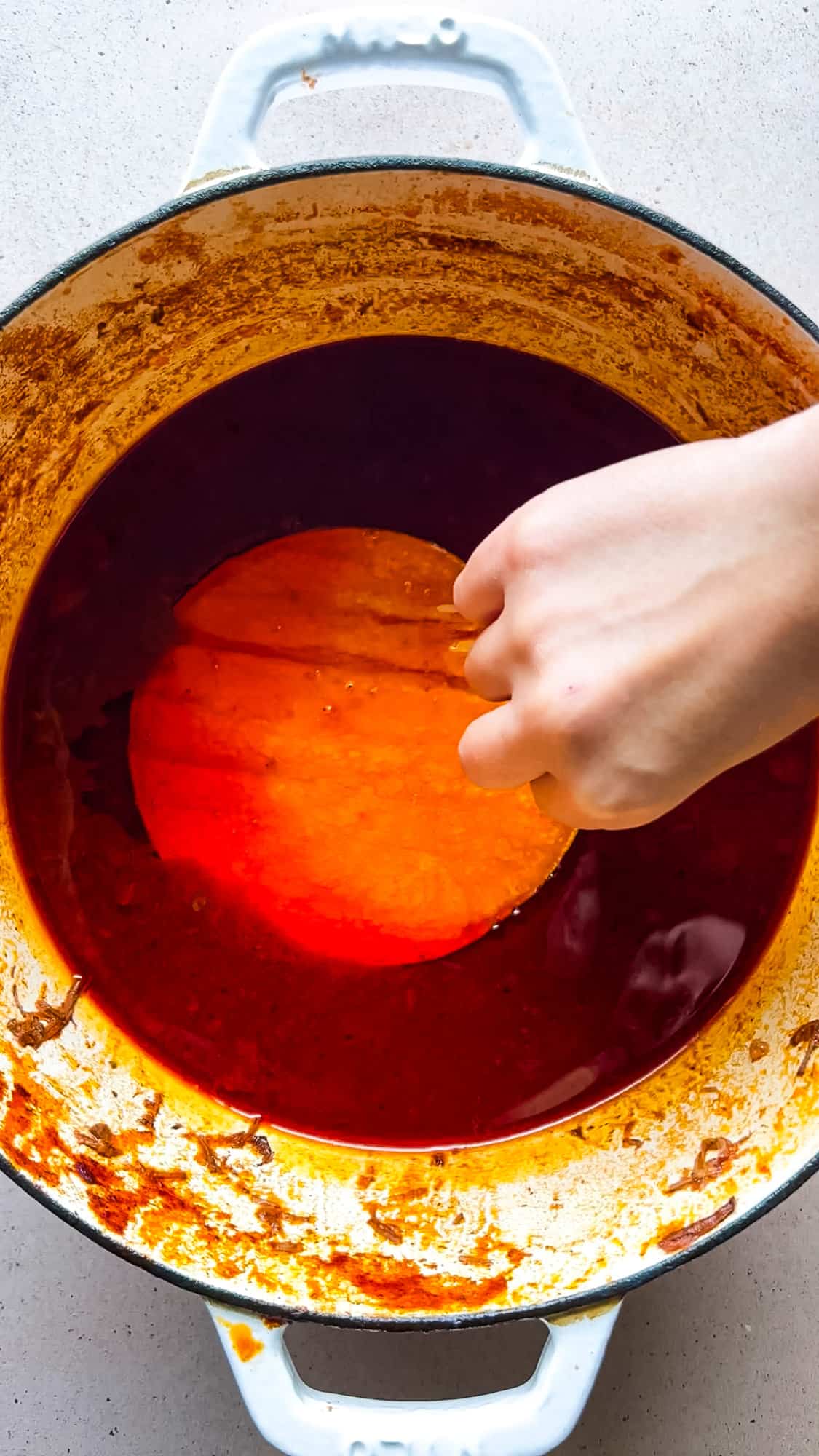 A corn tortilla being dipped into the top layer of fat of a pot of birria for making birria tacos.