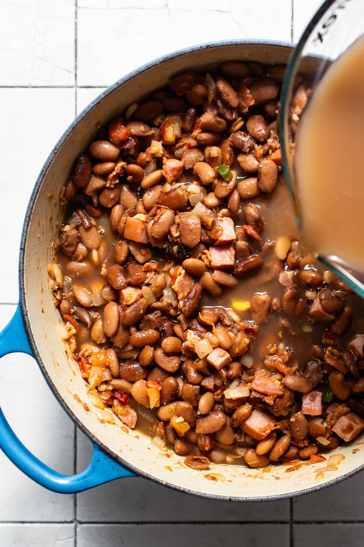 Pinto bean cooking liquid being poured into a pot of charro beans.