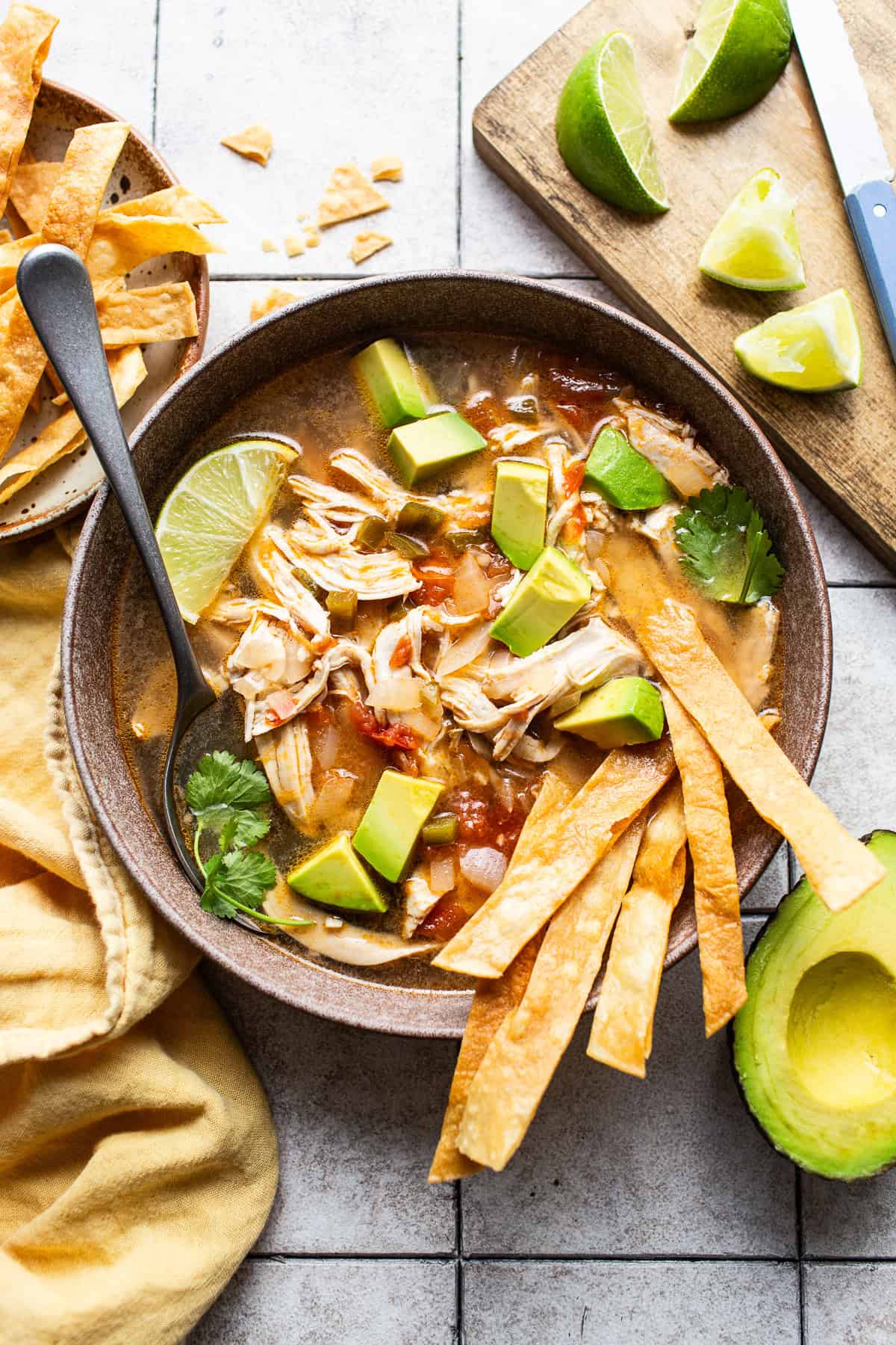 Sopa de lima served in a bowl with limes, diced avocados and tortilla strips. 