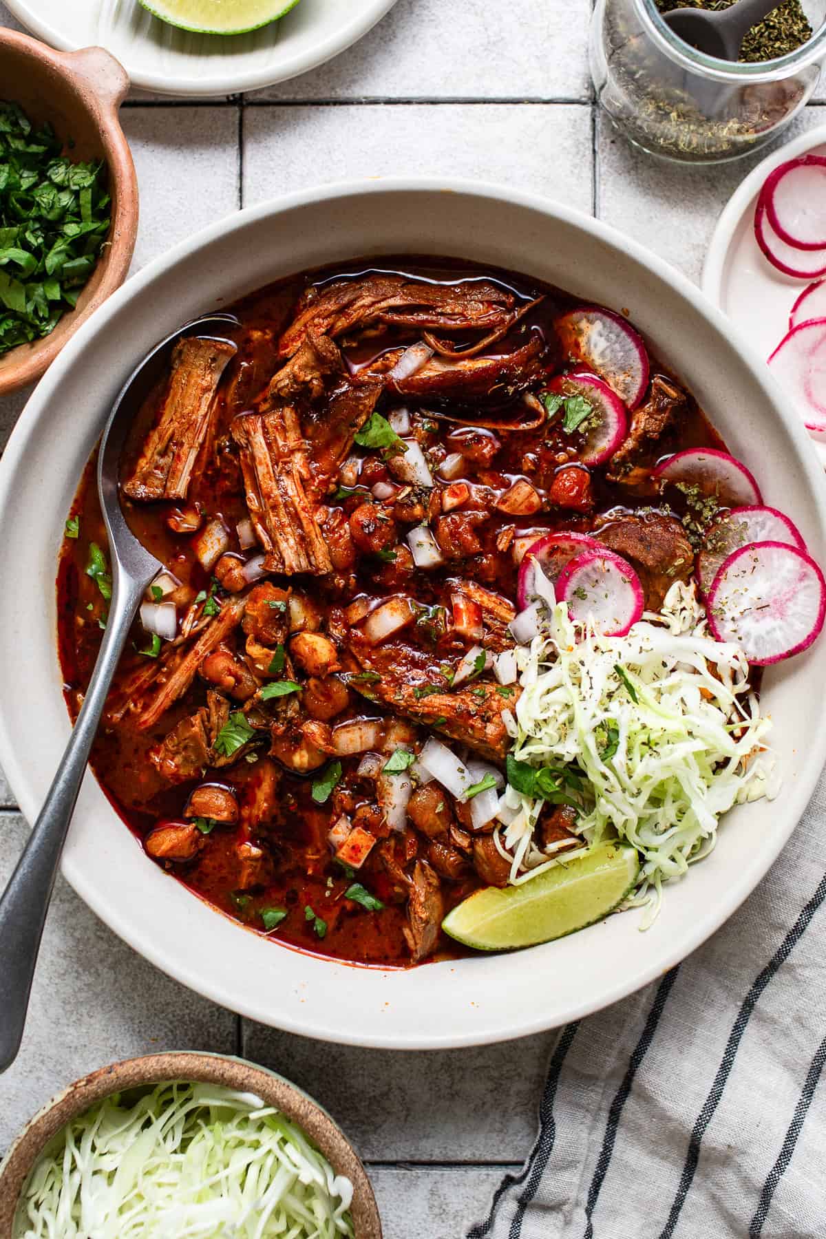 A large bowl of pozole rojo served with limes, oregano, radish slices, cilantro, and cabbage.