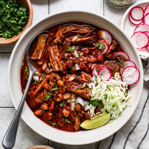 Pozole rojo in a bowl topped with shredded cabbage, onion, radishes, cilantro, and limes.
