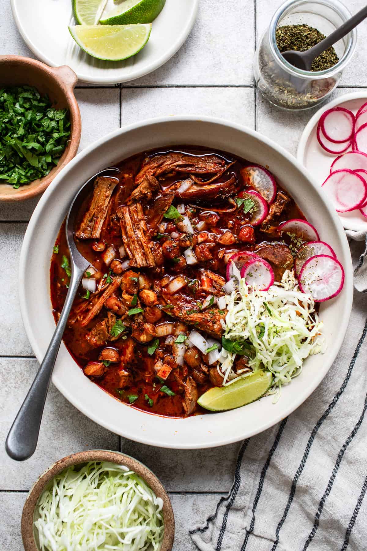 A large bowl of pozole rojo served with limes, oregano, radish slices, cilantro, and cabbage.