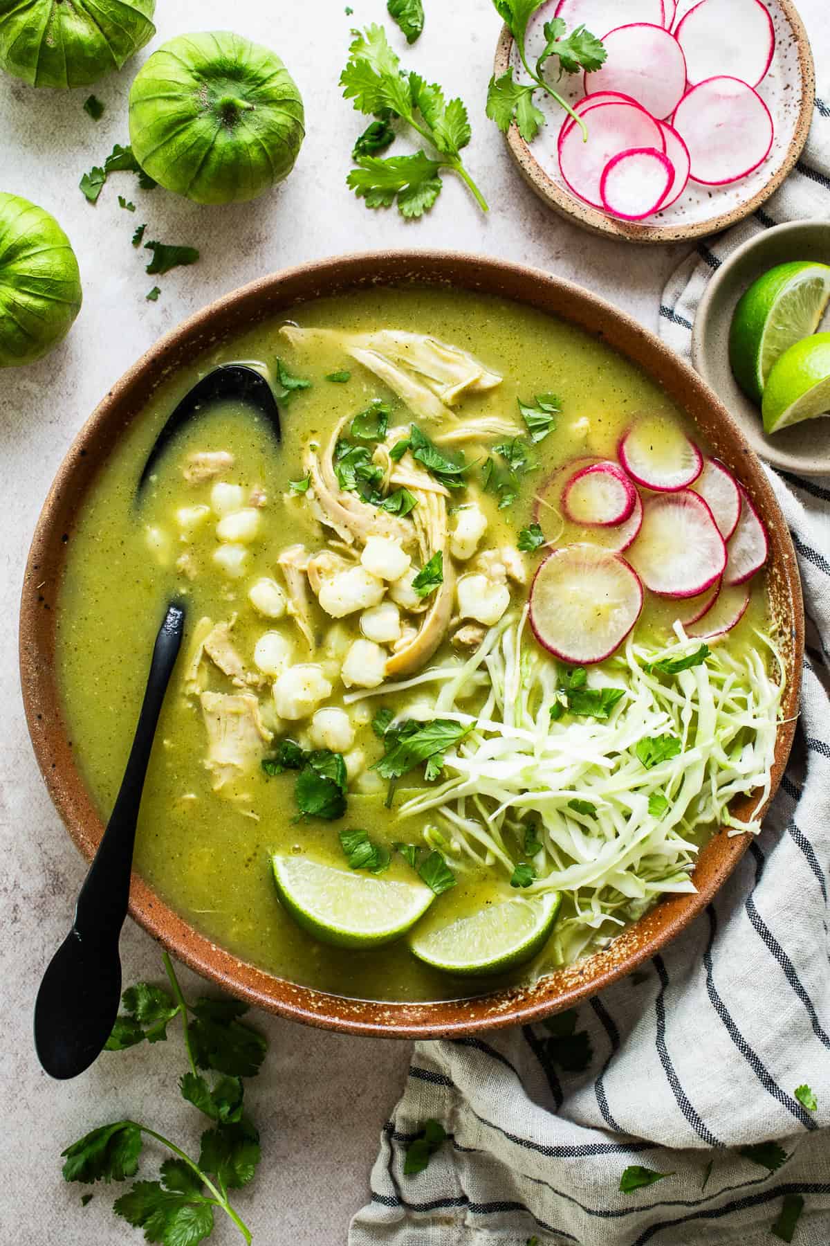 A bowl of pozole verde topped with shredded cabbage, radishes, cilantro, and served with fresh lime wedges.