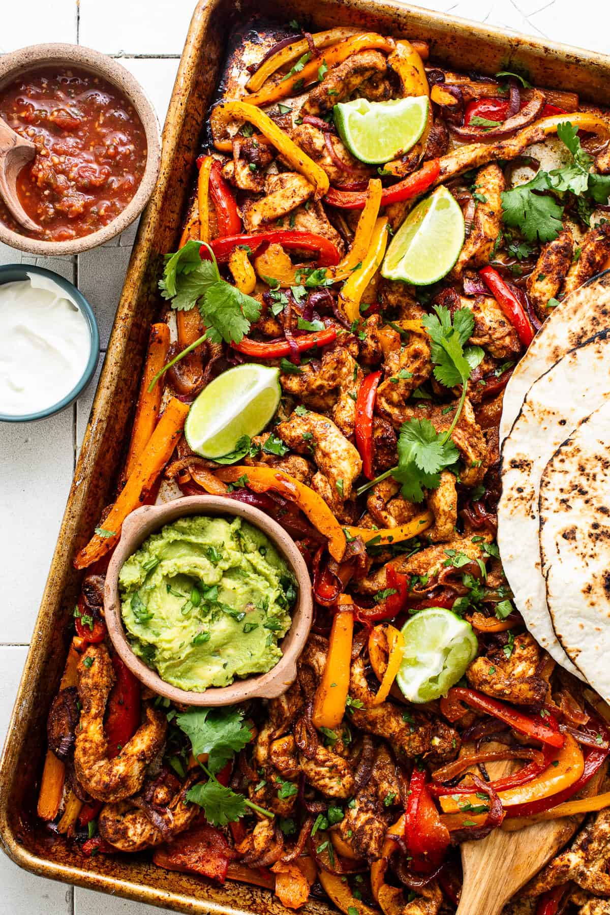 A bowl of guacamole and flour tortillas served with chicken fajitas on a sheet pan.