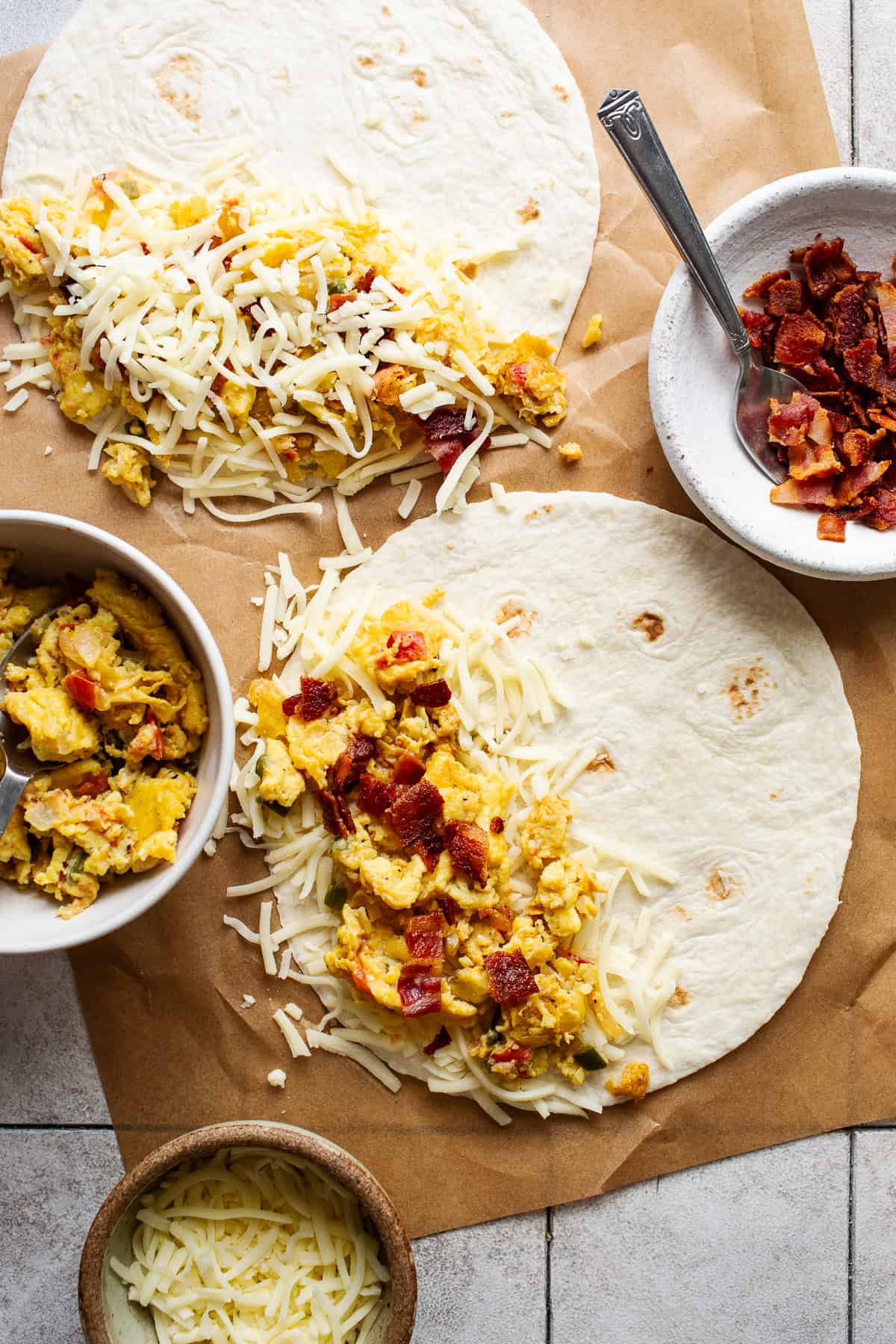 Breakfast quesadillas being assembled with scrambled eggs, shredded cheese, and crumbled cooked bacon.