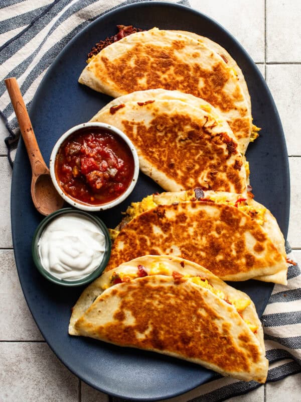 Four crispy and golden breakfast quesadillas on a plate served with salsa and sour cream.