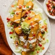 Bean and cheese taquitos on a large plate on top of shredded lettuce and served with a drizzle of Mexican crema, diced tomatoes, guacamole, and cotija cheese.
