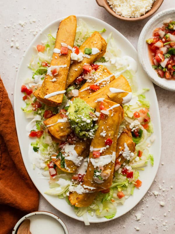 Bean and cheese taquitos on a large plate on top of shredded lettuce and served with a drizzle of Mexican crema, diced tomatoes, guacamole, and cotija cheese.