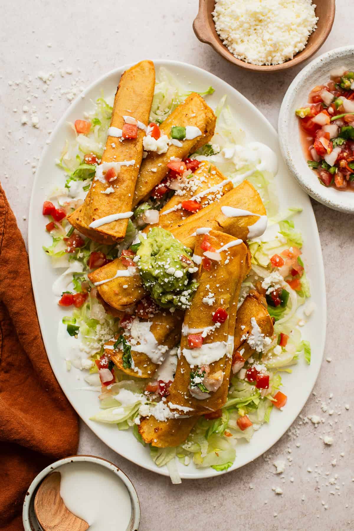 Bean and cheese taquitos on a large plate on top of shredded lettuce and served with a drizzle of Mexican crema, diced tomatoes, guacamole, and cotija cheese.