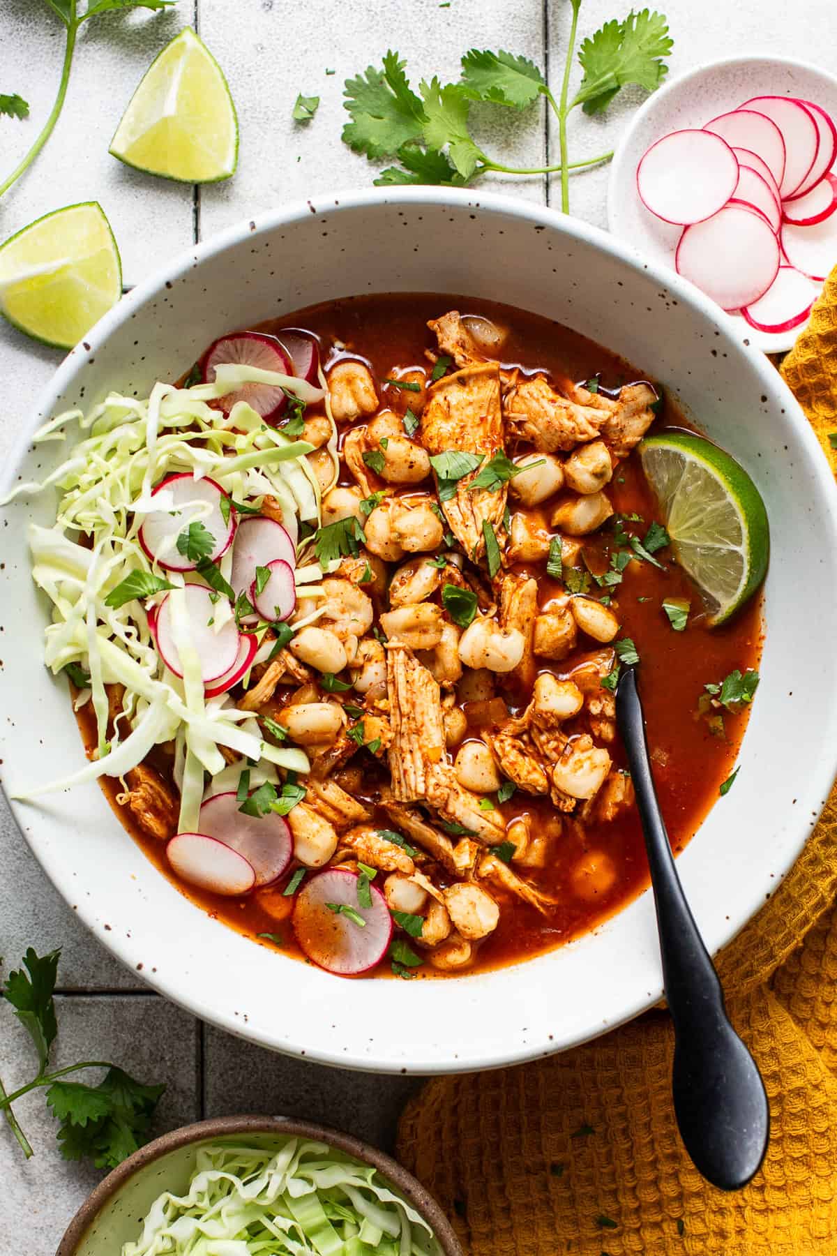 A bowl of easy chicken pozole topped with shredded cabbage, thinly sliced radishes, Mexican oregano, chopped cilantro, and lime wedges.