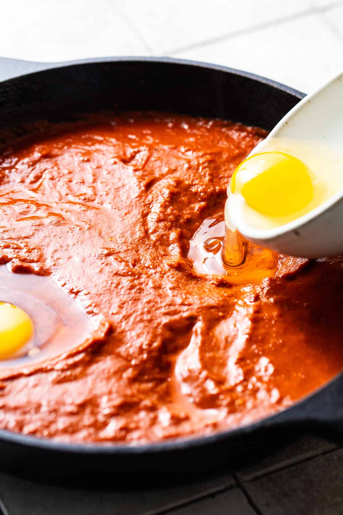 A cracked egg being poured into the sauce for huevos ahogados.