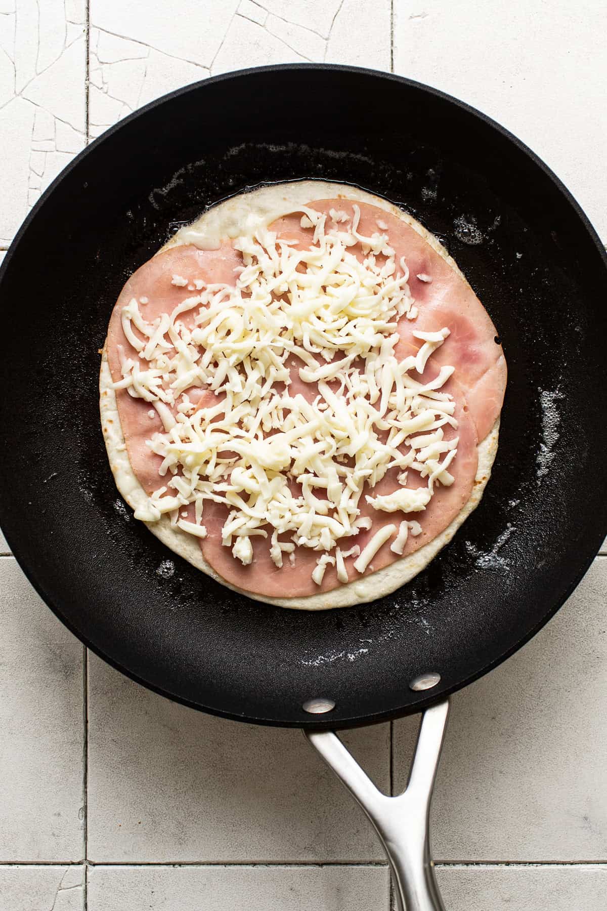 Sliced ham and shredded Mexican cheese on a flour tortilla on a skillet.