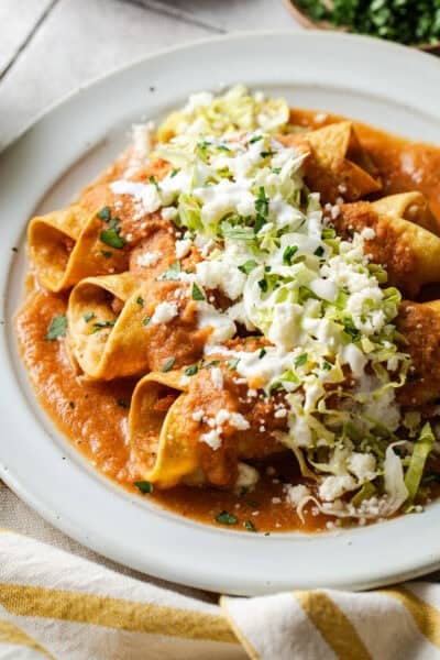 A plate of flautas ahogadas drowned in a salsa roja with shredded lettuce, crema, cotija cheese, and cilantro.