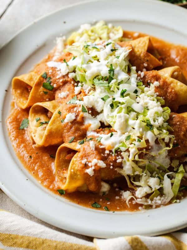 A plate of flautas ahogadas drowned in a salsa roja with shredded lettuce, crema, cotija cheese, and cilantro.