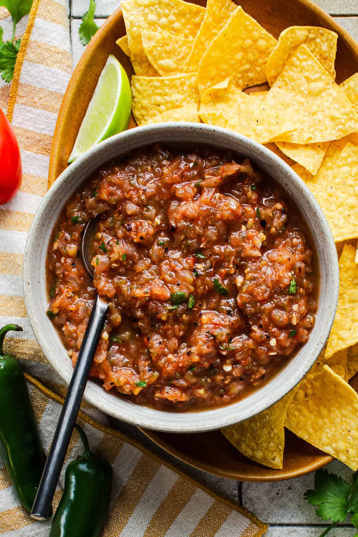 A bowl of roasted salsa made with fresh tomatoes on a platter next to tortilla chips.
