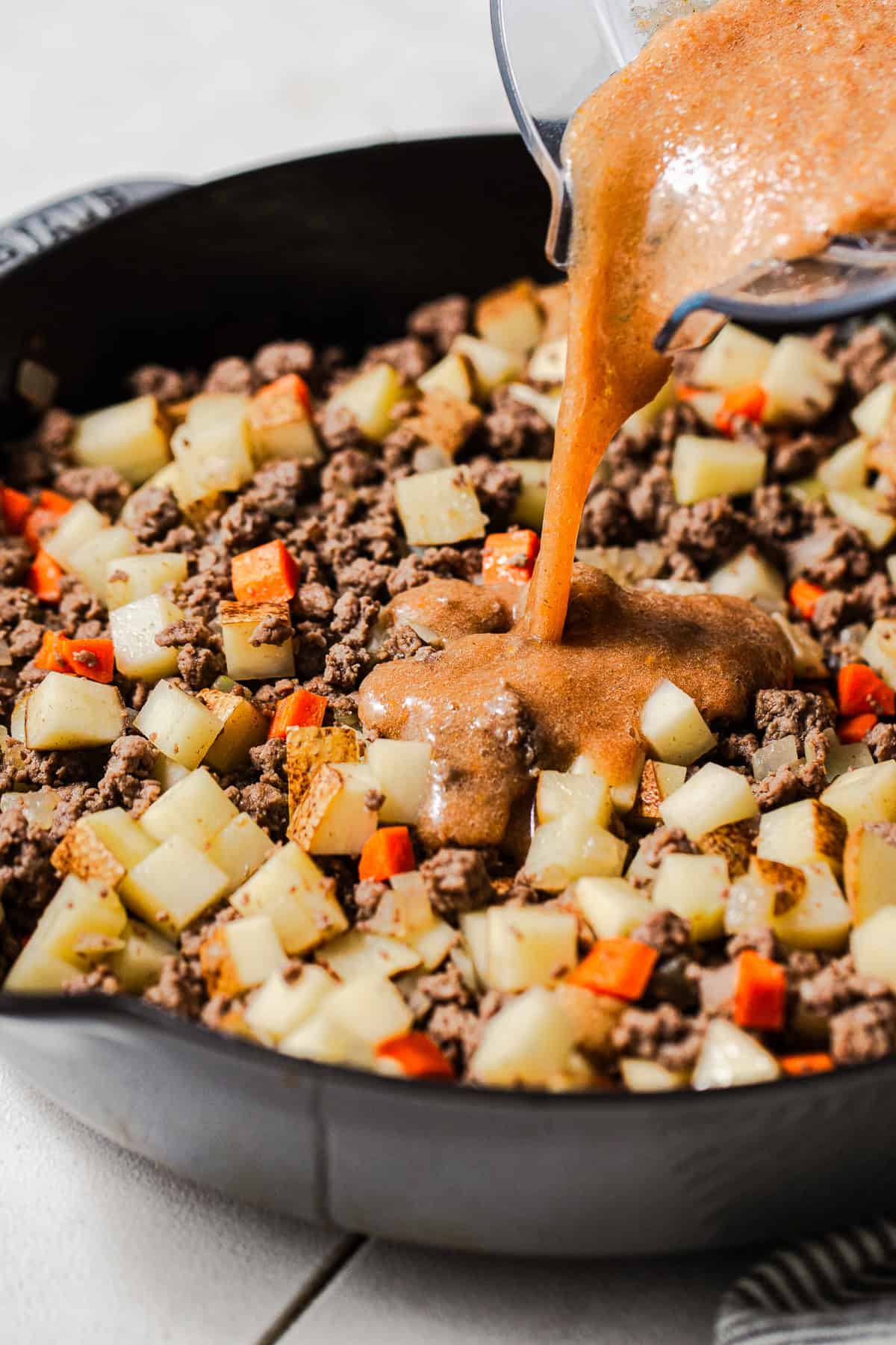 Blended tomato sauce being poured into the skillet of beef picadillo.