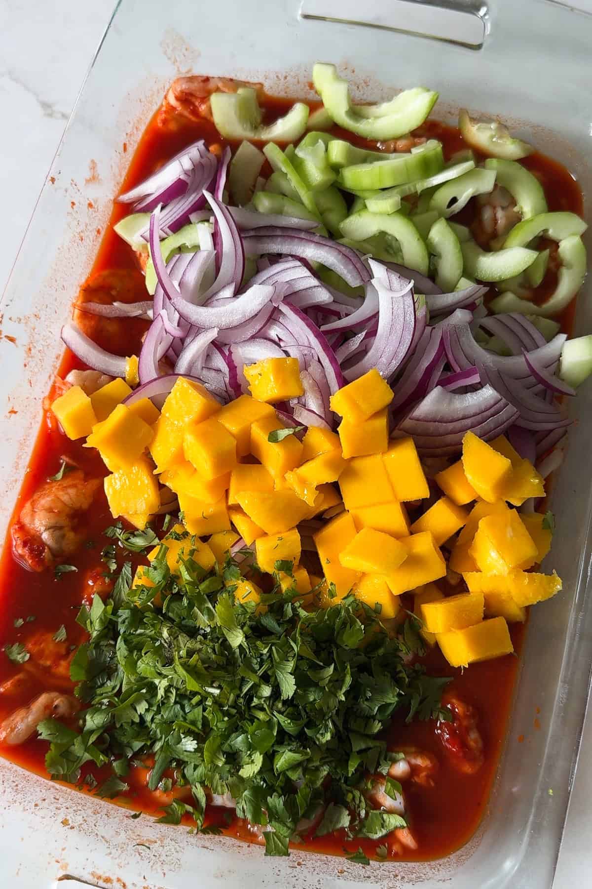 Cucumber, red onion, mango, and cilantro on top of a dish full of aguachile rojo before being mixed in.