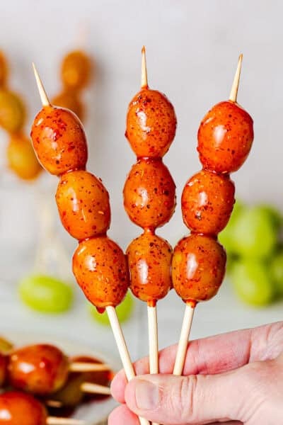 Hand holding three skewers of boozy grapes, aka tequila grapes, covered in chamoy and Tajín (chile lime seasoning).