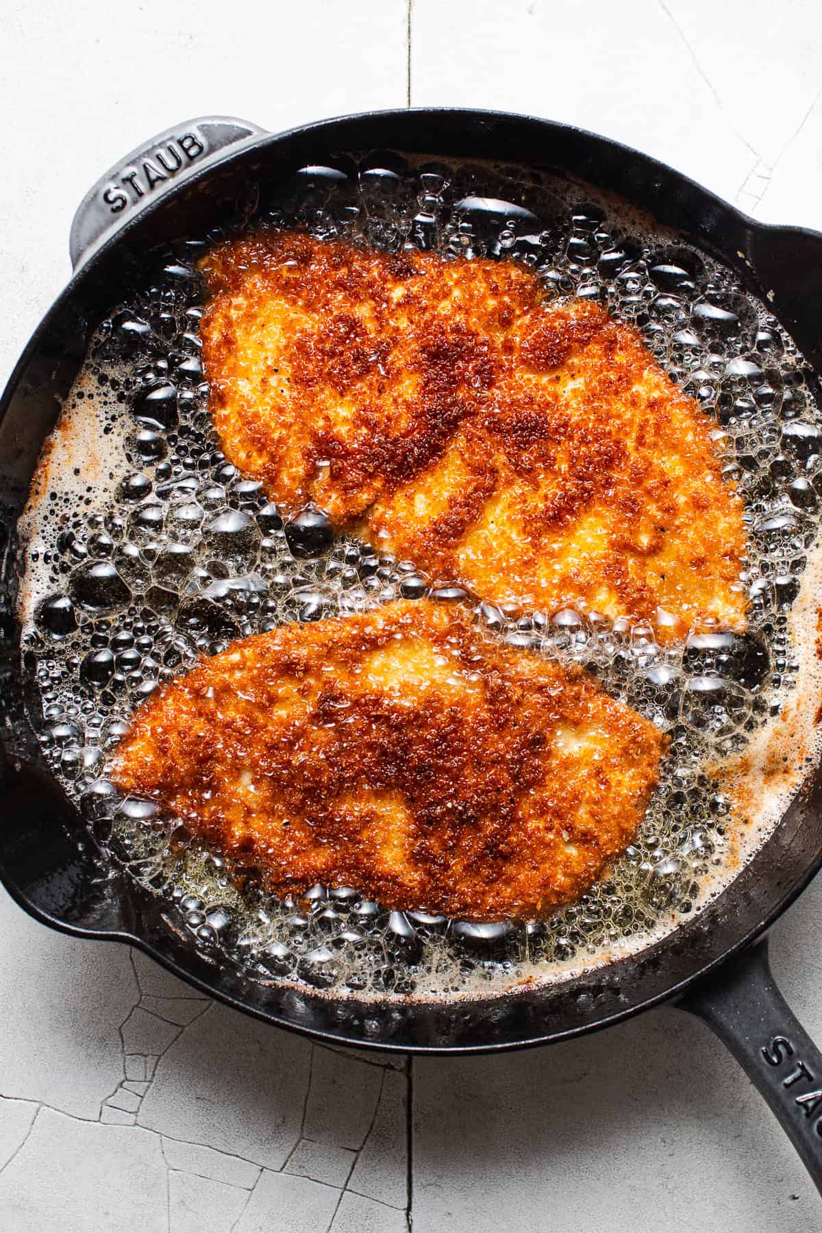 Two chicken milanesa cutlets frying in a skillet.