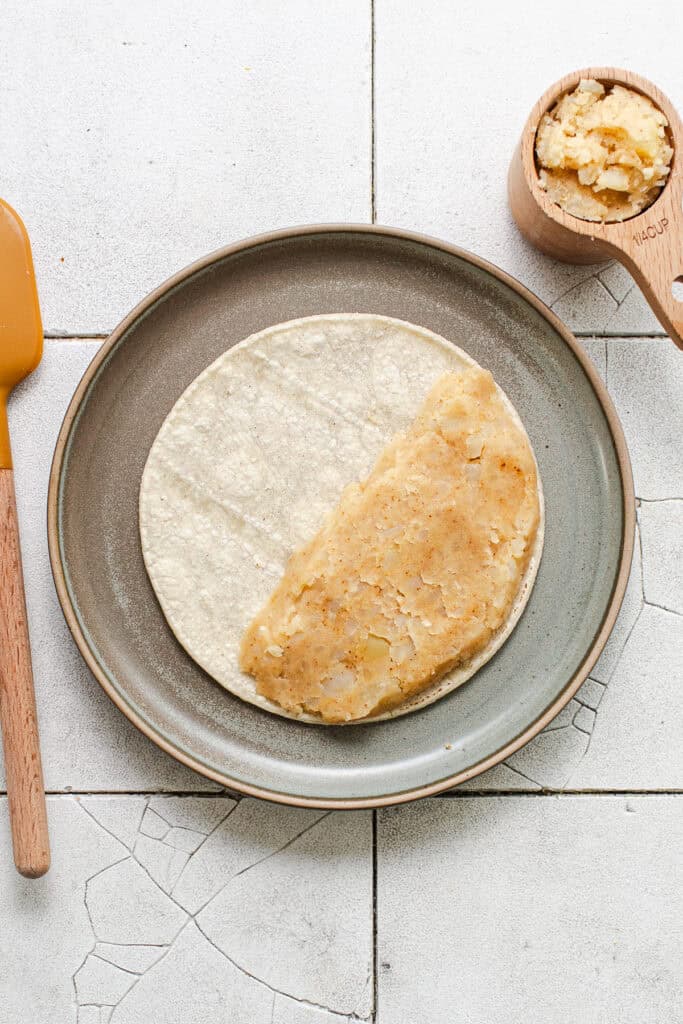 A potato taco with the seasoned mashed potato filling on one side of a tortilla before being folded and fried.